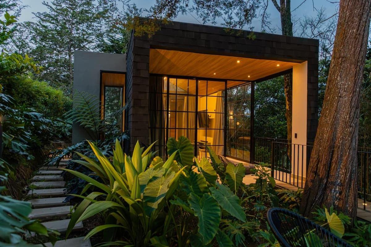 Fachada de cabaña moderna con grandes ventanales iluminados rodeada de vegetación tropical en Rionegro Antioquia