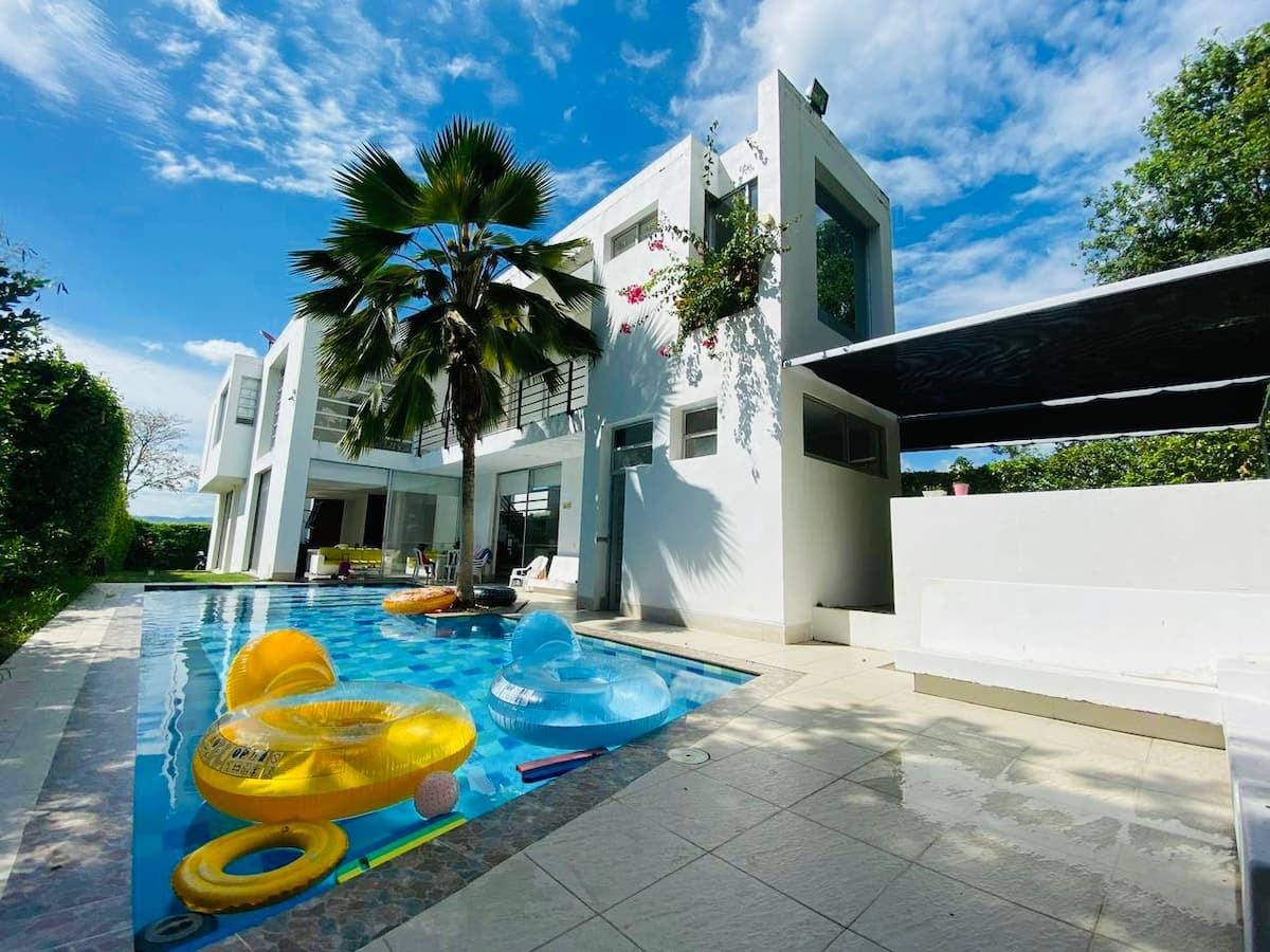 Casa moderna con piscina azul y flotadores amarillos en Apulo, rodeada de palmeras tropicales